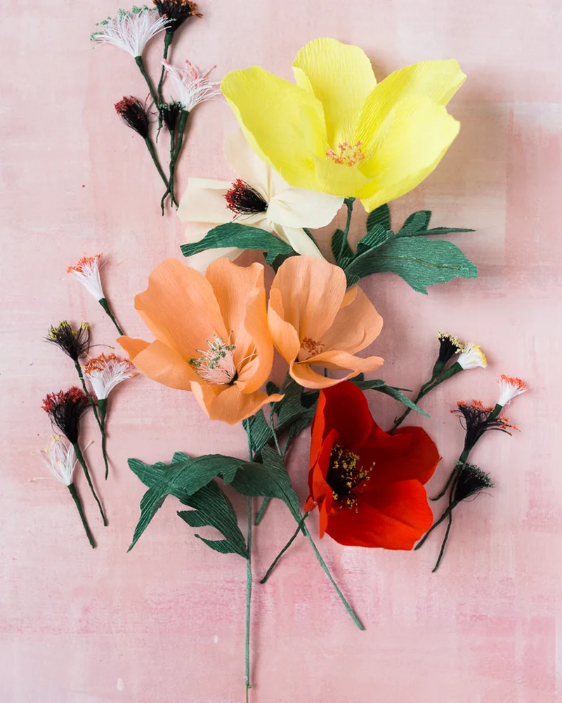 Crepe paper poppies and wildflowers with dark stamens laid flat on a pink background