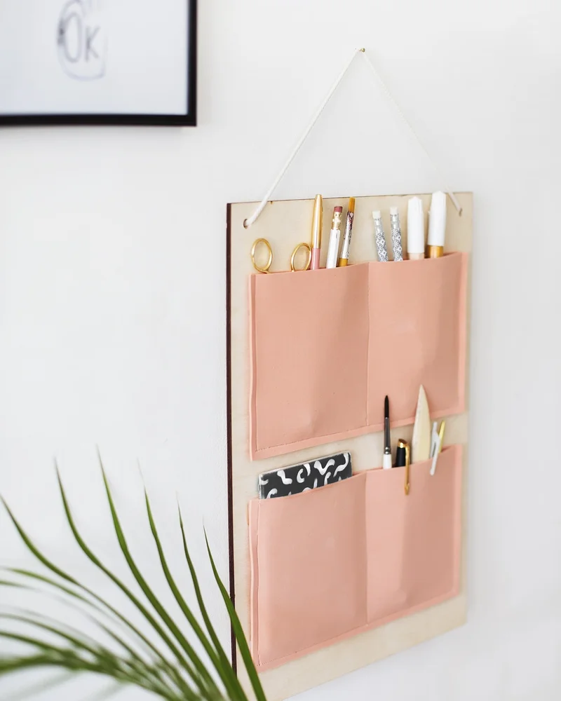 Wood board wall organizer with blush pink fabric pockets holding pens and scissors