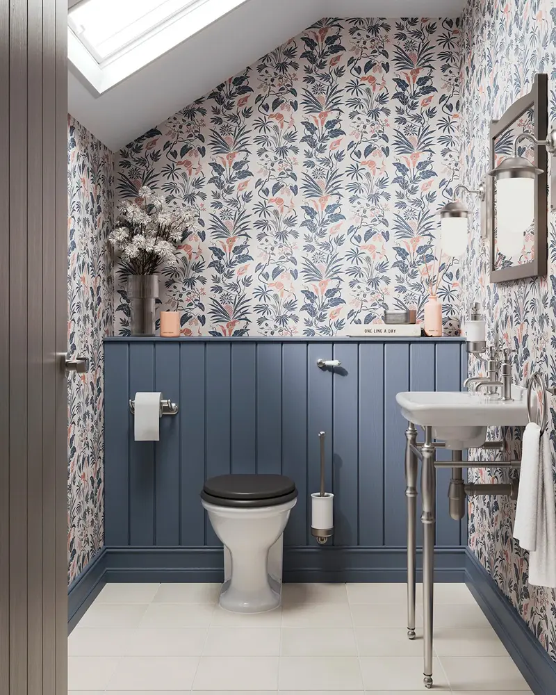 Small bathroom with slate blue beadboard wainscoting below blue and pink floral wallpaper under a sloped ceiling