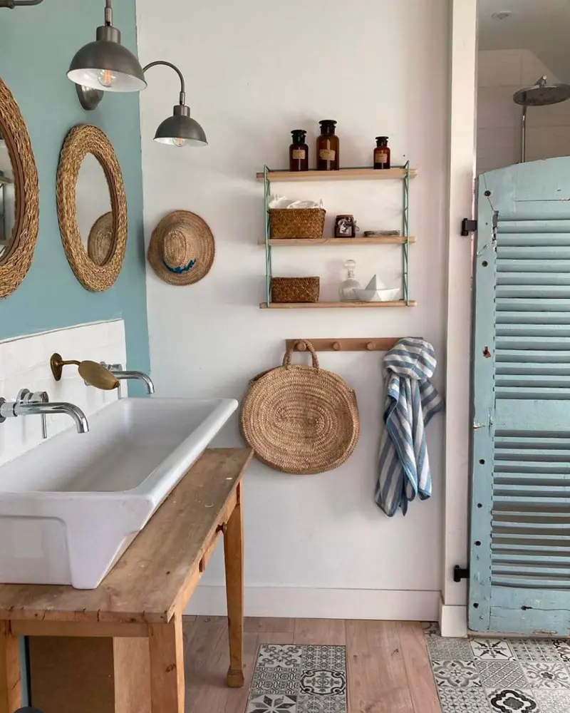 Bathroom with a soft blue wall, a white trough sink on a wooden table, rattan mirrors, a woven bag and a blue louvered shutter