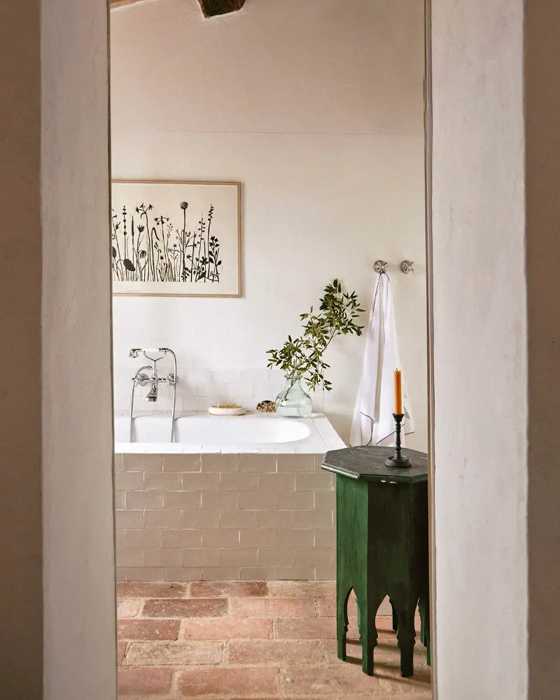 View through a doorway to a tiled tub, a brick-paved floor, a green carved side table and a botanical print