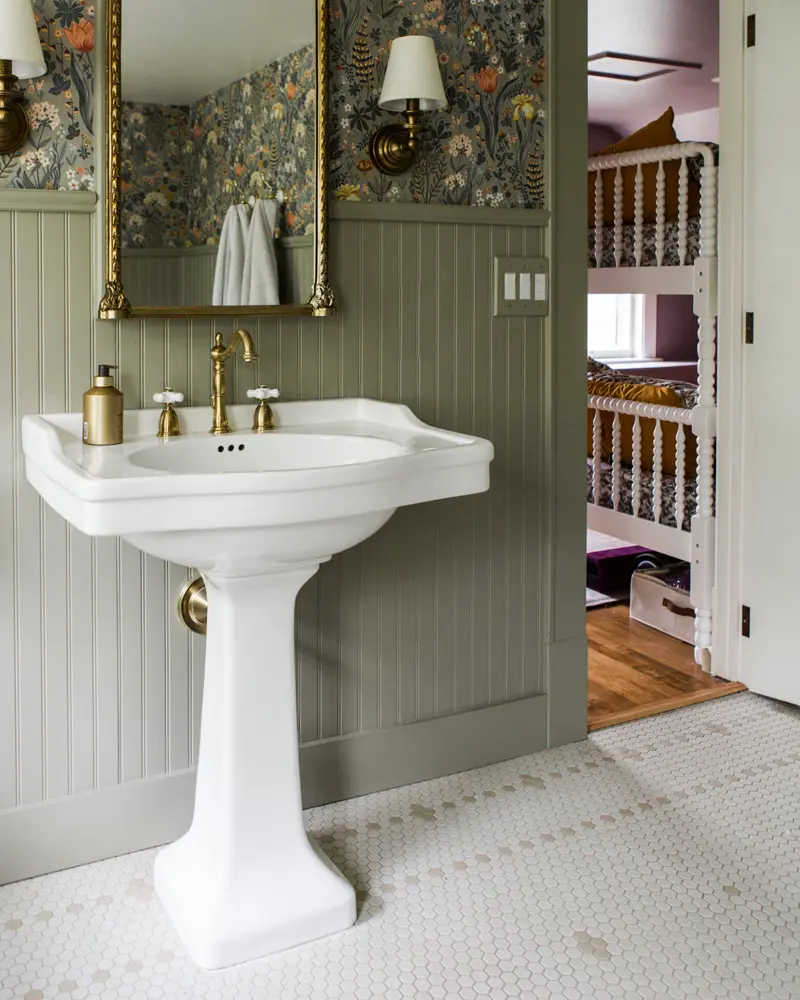 White pedestal sink with brass faucets, green paneled wainscoting, a botanical wallpaper and a gilded mirror