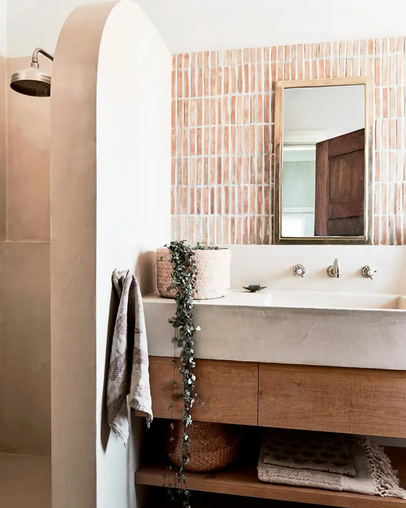 Pink vertical tiles behind a concrete trough sink on a wood vanity with towels on open shelving under an arched doorway