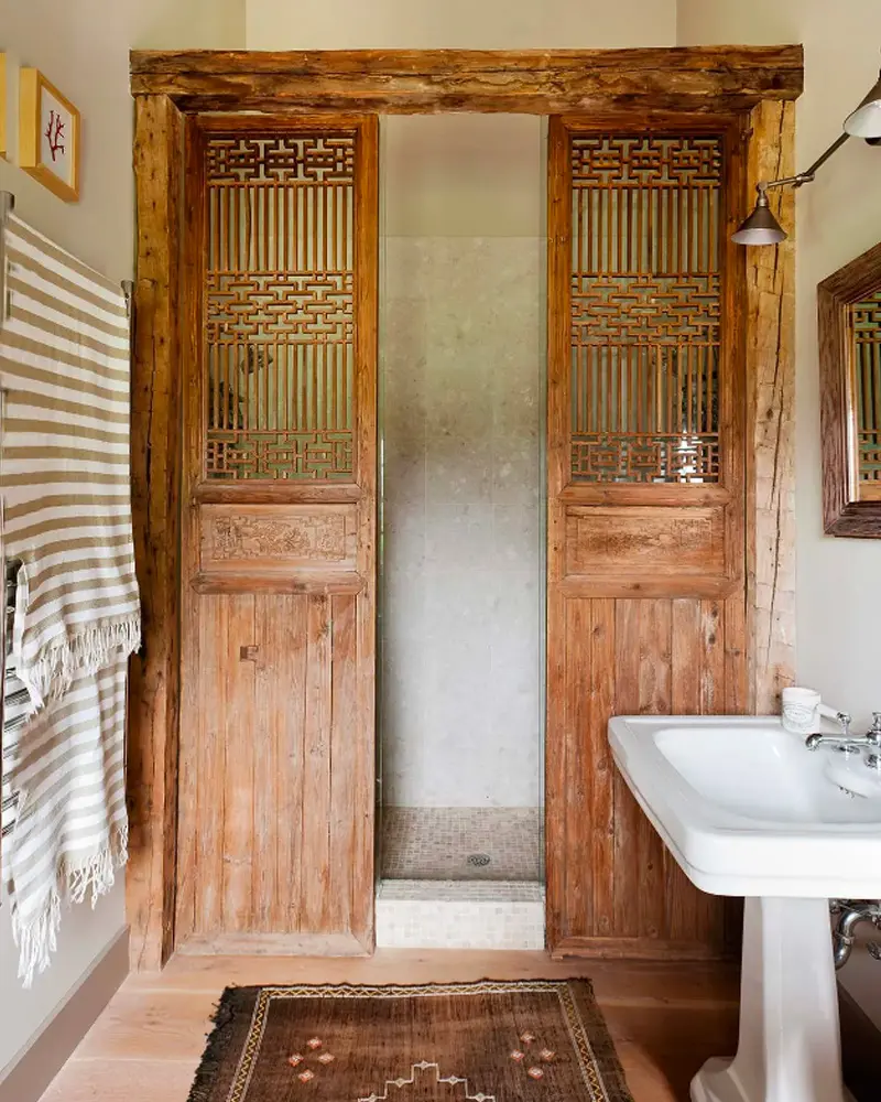 Pair of antique carved wooden doors framing a walk-in shower beside a white pedestal sink