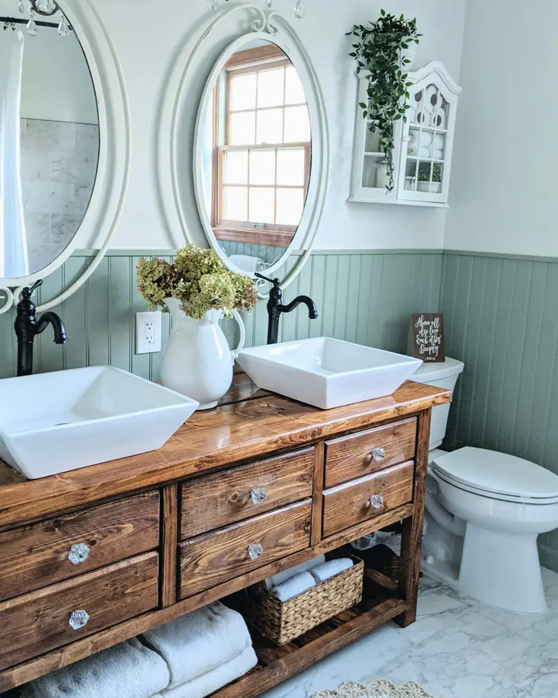 Double vanity made from rustic wood drawers with two white square sinks, oval white mirrors and sage green beadboard