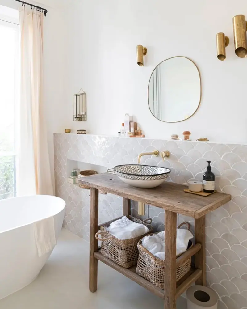 Minimalist bathroom with white scallop tiles, brass sconces, a round mirror and a rustic wood washstand with a patterned vessel sink