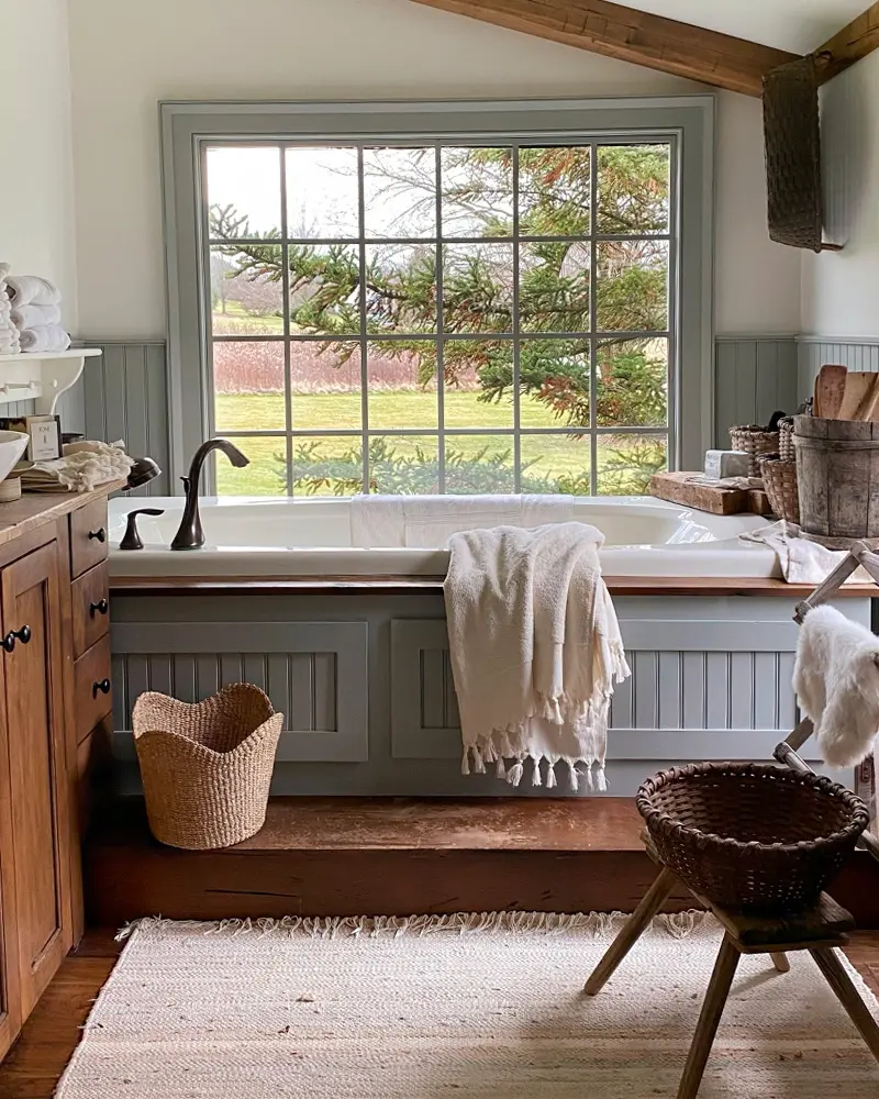 Built-in soaking tub with sage gray paneling under a large picture window overlooking trees, with a wood vanity and baskets