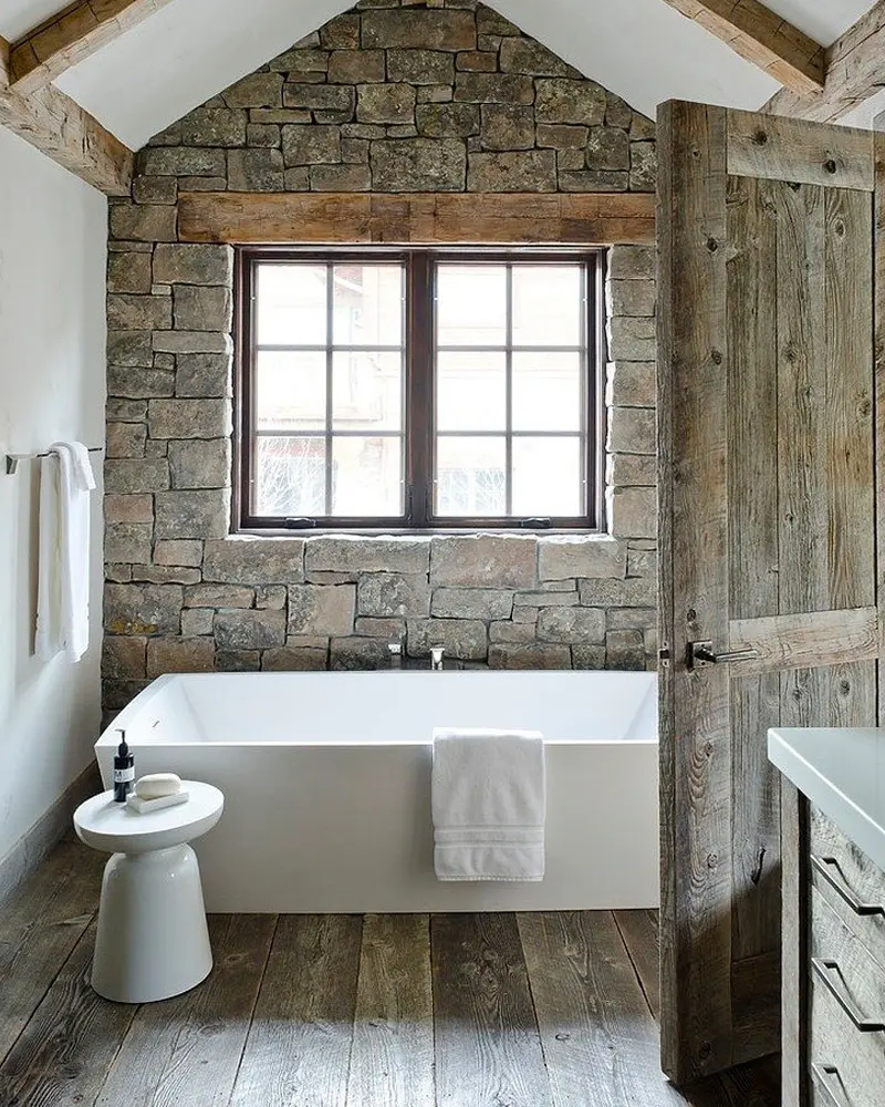 Bathroom with a fieldstone accent wall around a window, a white rectangular tub, a wide-plank floor and a rustic barn door
