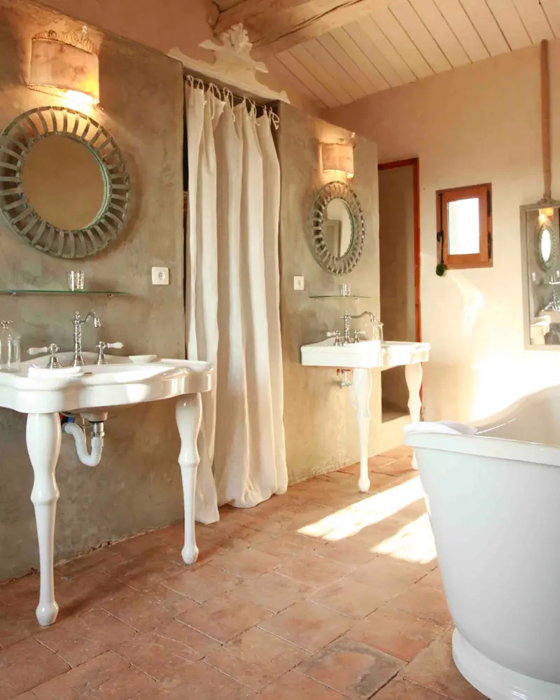 Two white console sinks with round mirrors, a terracotta floor, a linen shower curtain and a freestanding tub under a wood ceiling