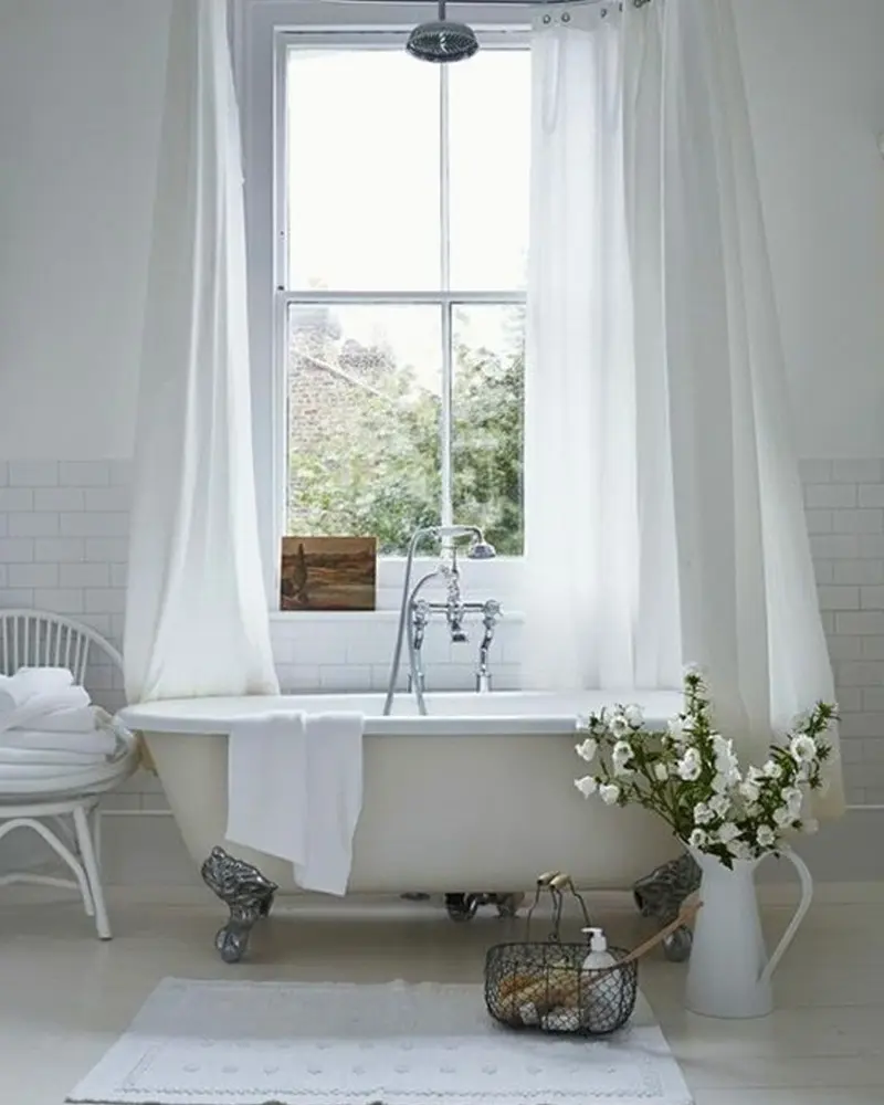 White clawfoot tub beneath a tall window with sheer white curtains, a wire basket and a jug of white flowers