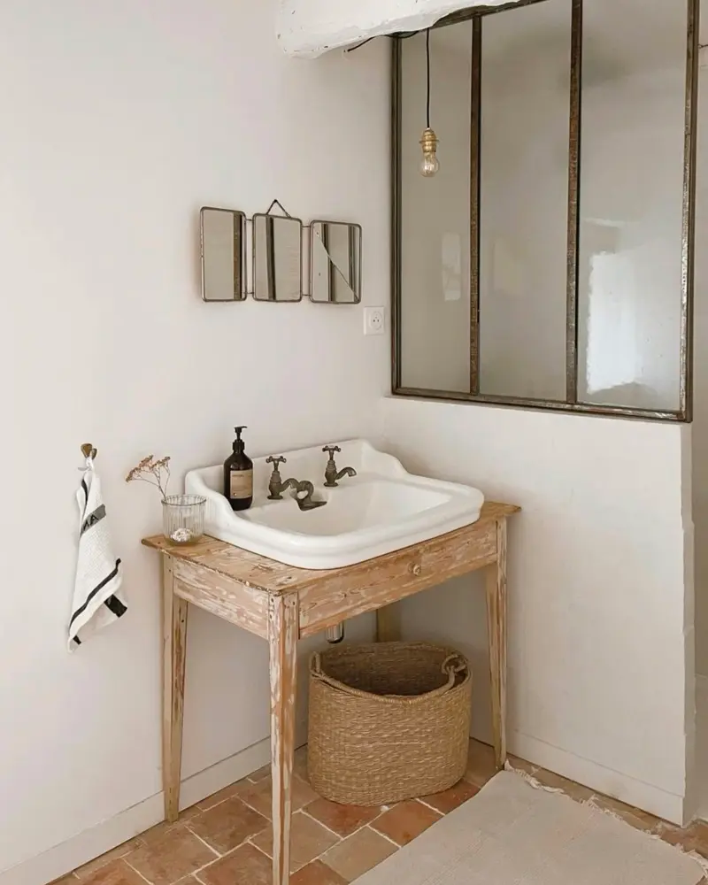 Whitewashed wooden washstand with a white sink and woven basket beside a steel-framed glass partition and terracotta floor