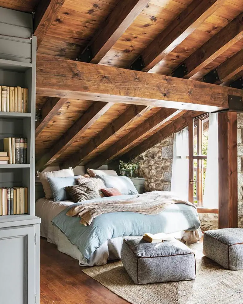 Attic bedroom with dark wood roof beams, a stone wall, blue bedding and knit poufs