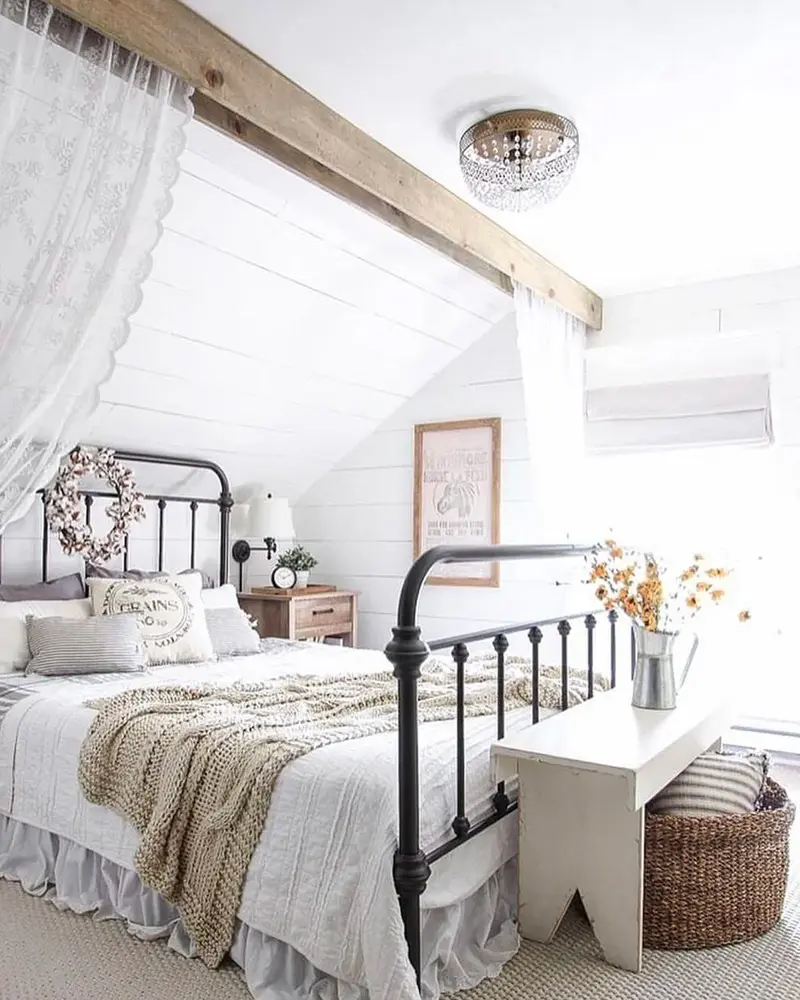 Black iron bed with white and cream bedding under a sloped white ceiling with a wood beam
