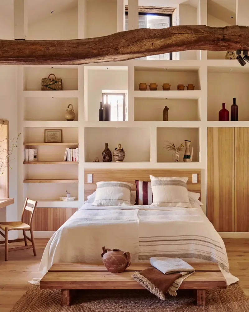 Live-edge wood beam above a bed backed by built-in white shelves with pottery