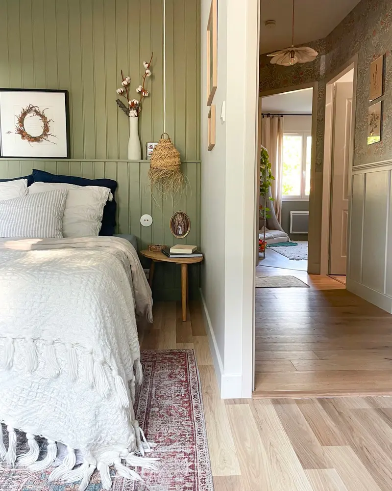 Sage green beadboard paneling behind a white bed with a red vintage runner on wood floors