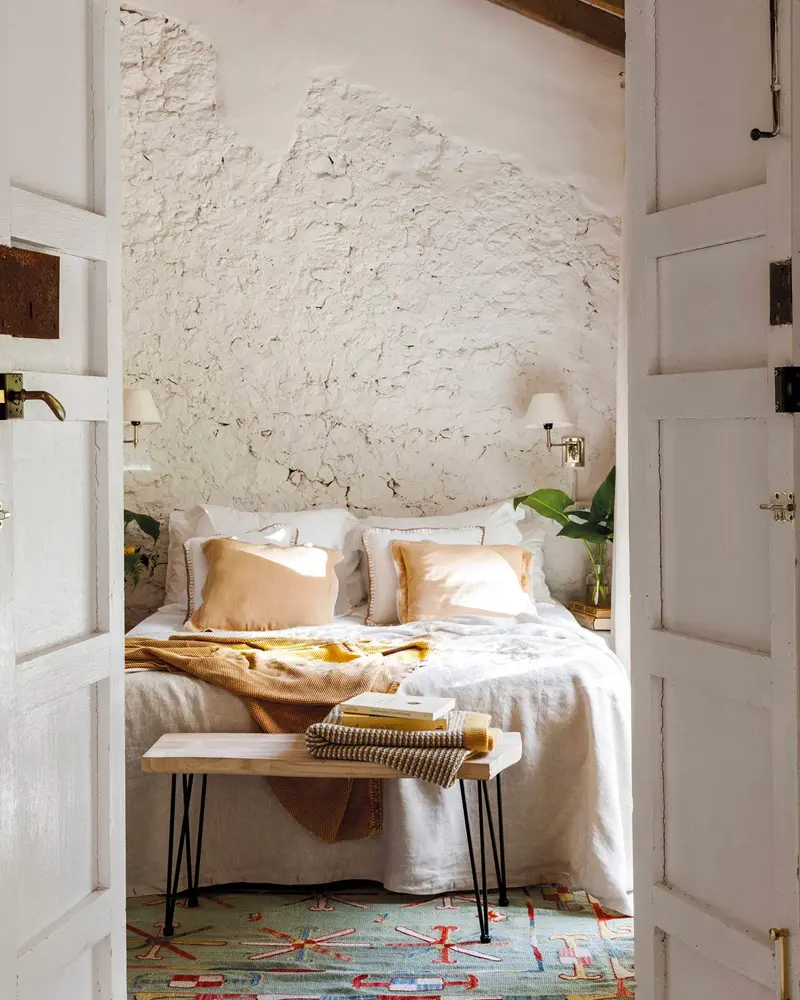 Whitewashed stone wall behind a bed with peach pillows, a hairpin-leg bench and a colorful rug