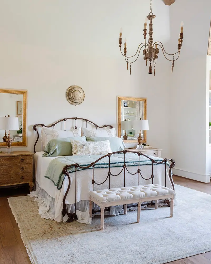 Scrolled iron bed, a wood bead chandelier, gilt mirrors and a tufted bench in a white room