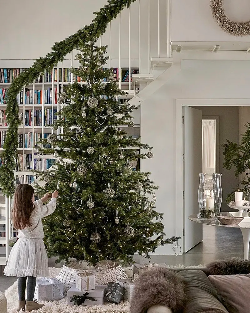 Girl decorating a tall tree with silver ornaments beside a staircase garland and bookshelves