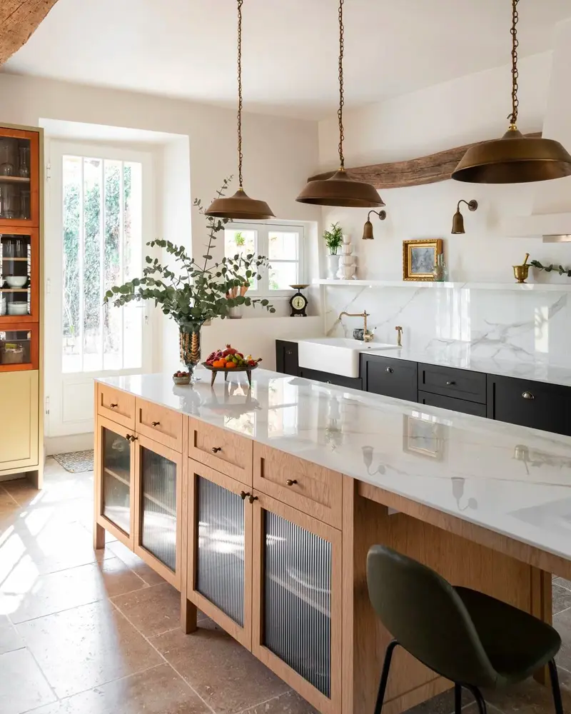 Long wood island with a white marble top and glass-front doors under brass pendants