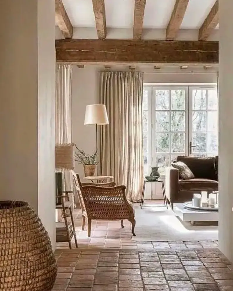 Terracotta floor tiles and dark beams leading to a sitting area with linen curtains and a wicker armchair