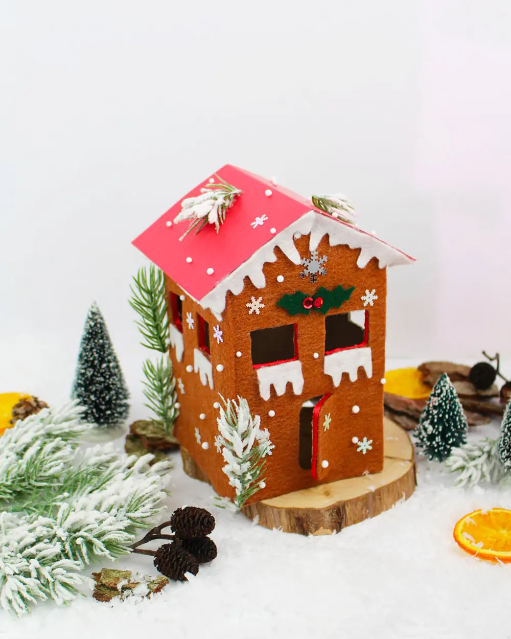 Brown felt gingerbread-style house with a red roof and white trim among mini trees and fake snow