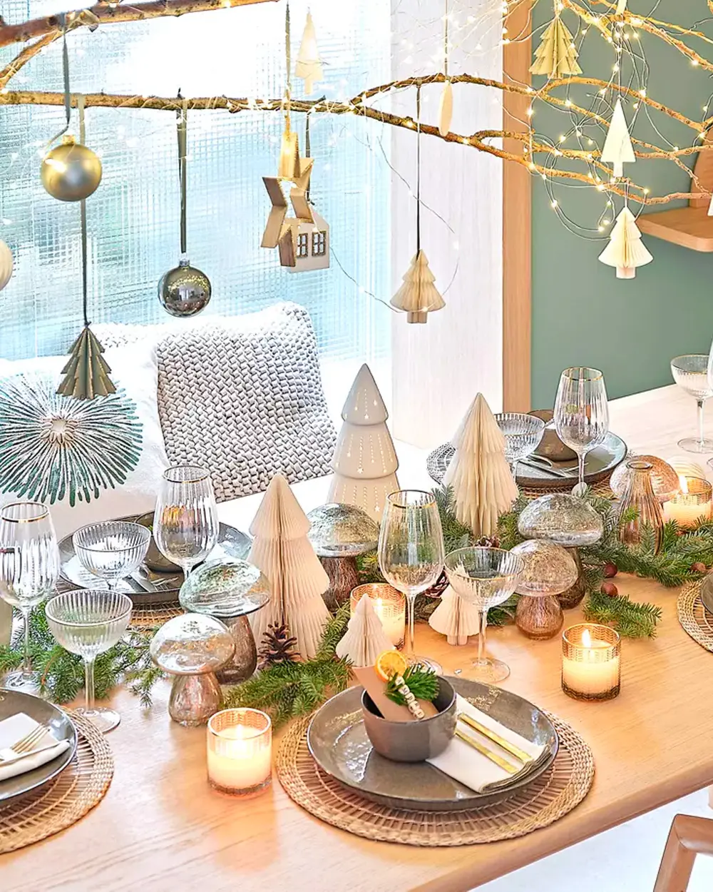 Light wood table with paper trees, glass mushrooms, greenery and paper ornaments hanging from a branch