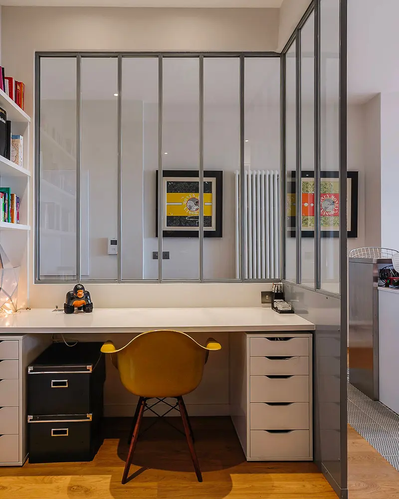 Black steel glass partition enclosing a desk with white drawers and yellow chair