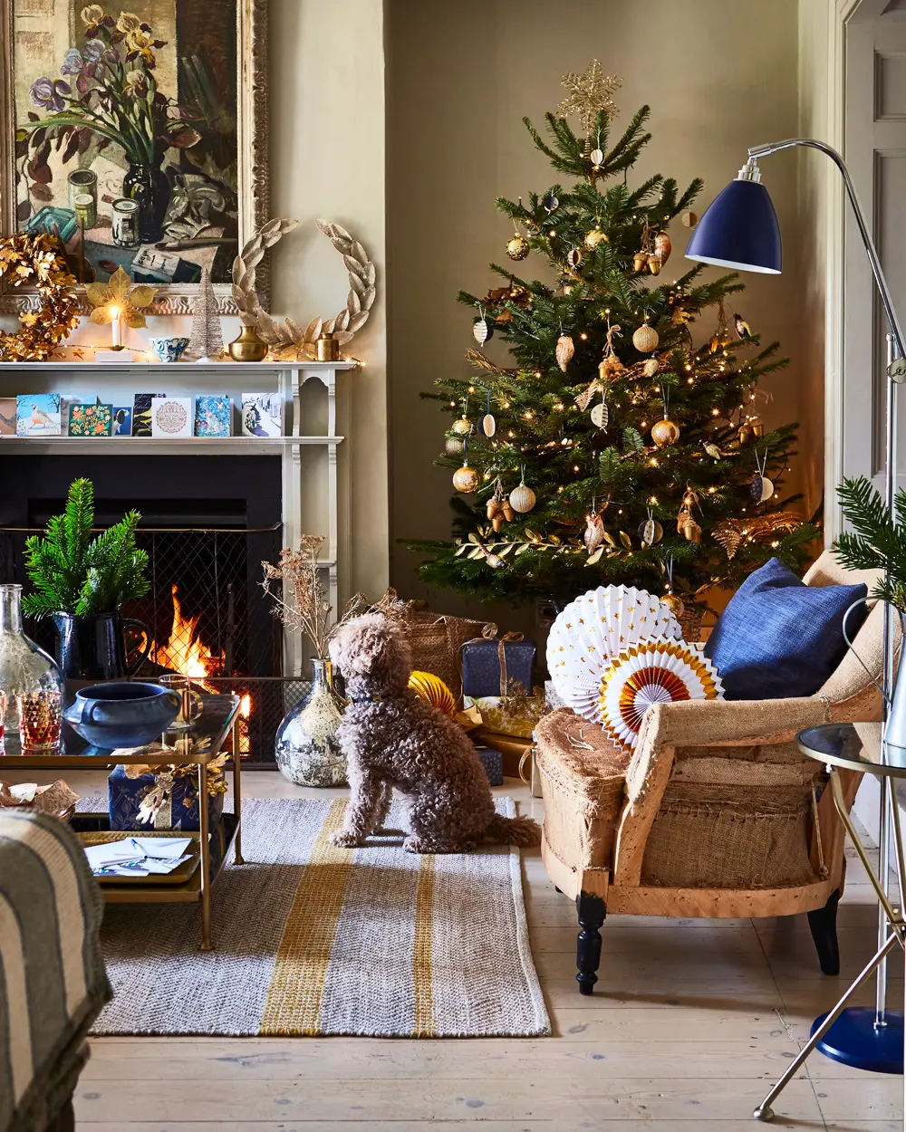 Living room with a gold-trimmed Christmas tree, navy pillows, a blue floor lamp and a dog by the fire