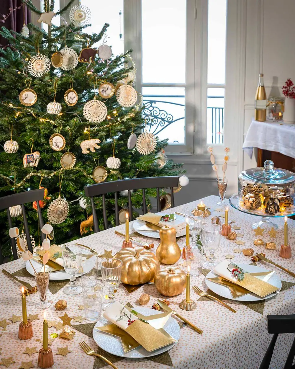 Dining table set with gold flatware and gold pumpkins beside a tree hung with paper medallions
