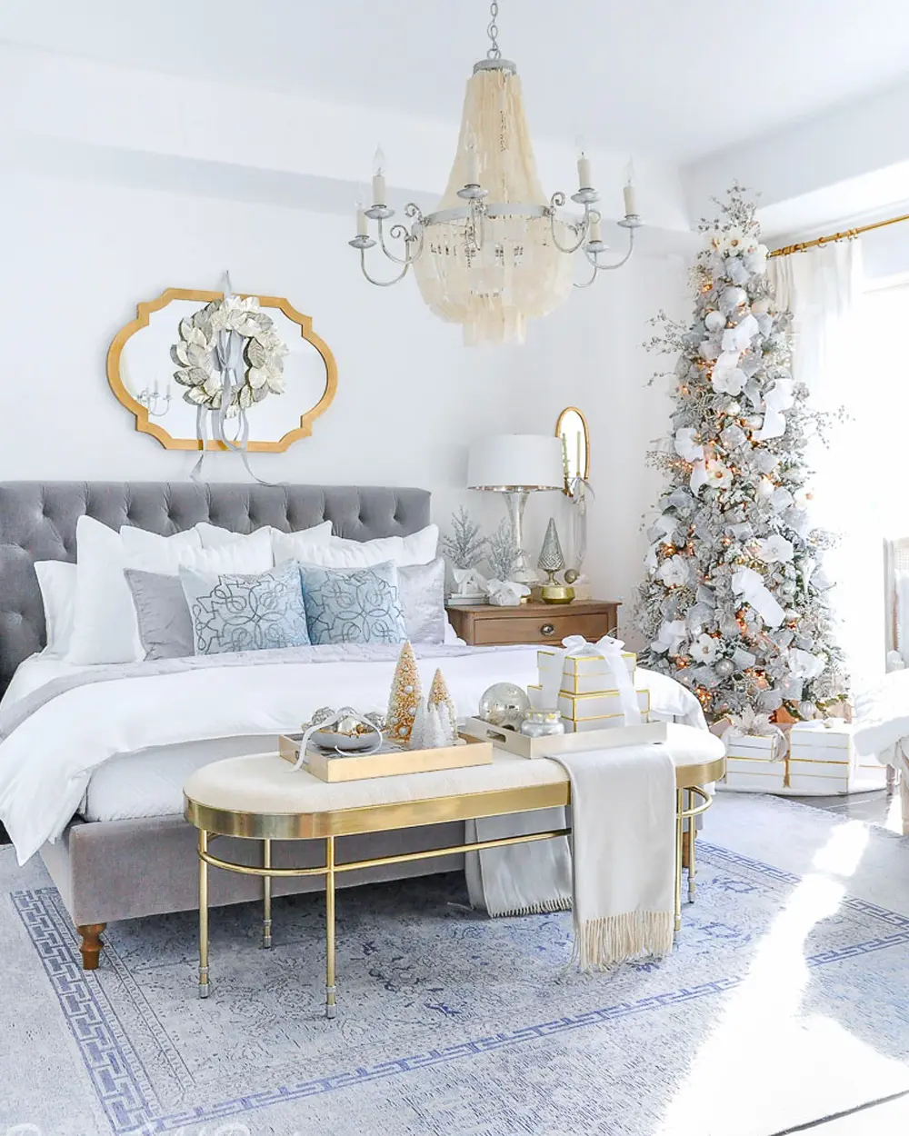 White bedroom with gray tufted bed, gold-framed mirror, gold bench and a white flocked tree