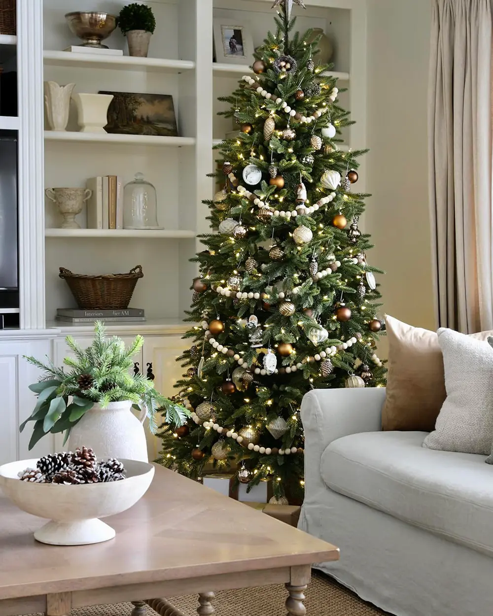 Tall green Christmas tree with gold and white ornaments and wood bead garland beside built-in shelves