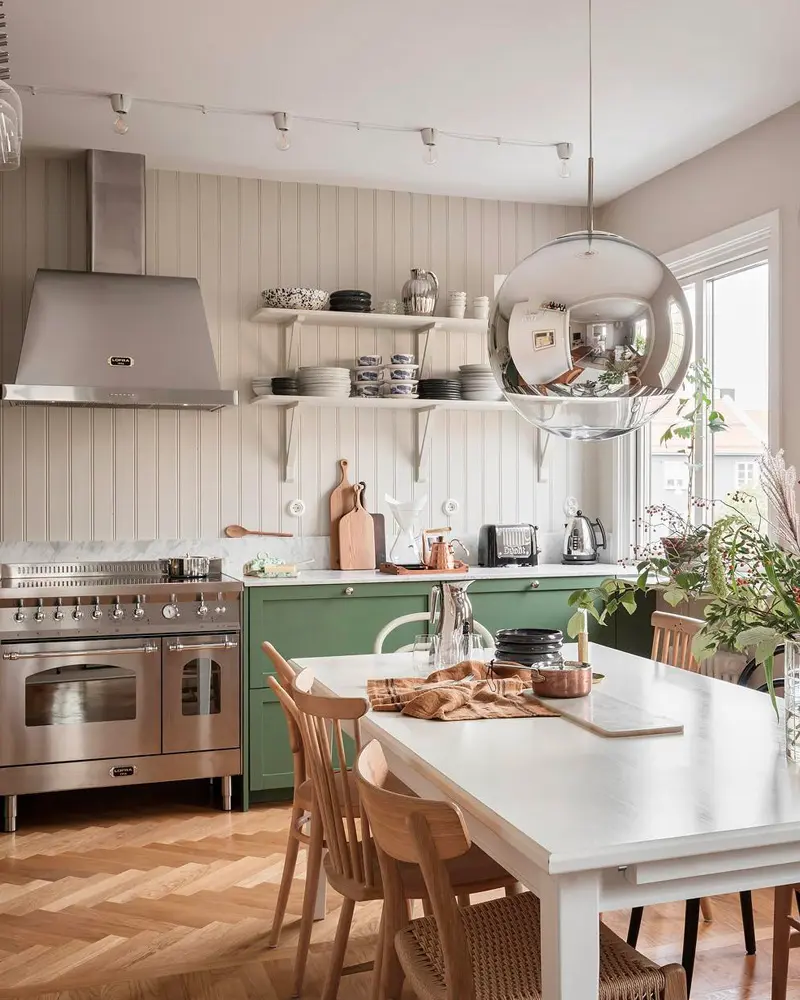 Green lower cabinets, stainless range, open shelves, chrome pendant and white table with wood chairs
