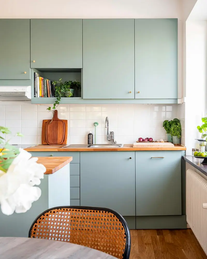 Sage green cabinets with wood counter, open cubby shelf and rattan-back chair at a round table