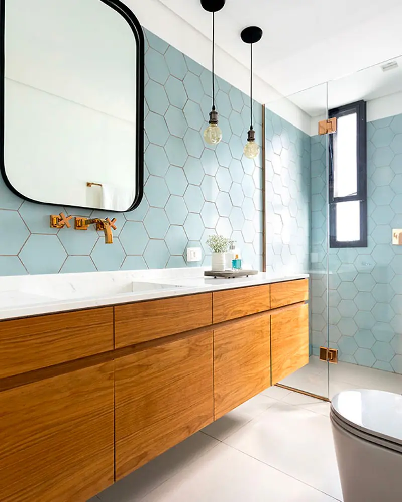 Long wood vanity under a black-framed mirror with pale blue hexagon wall tiles and brass faucet