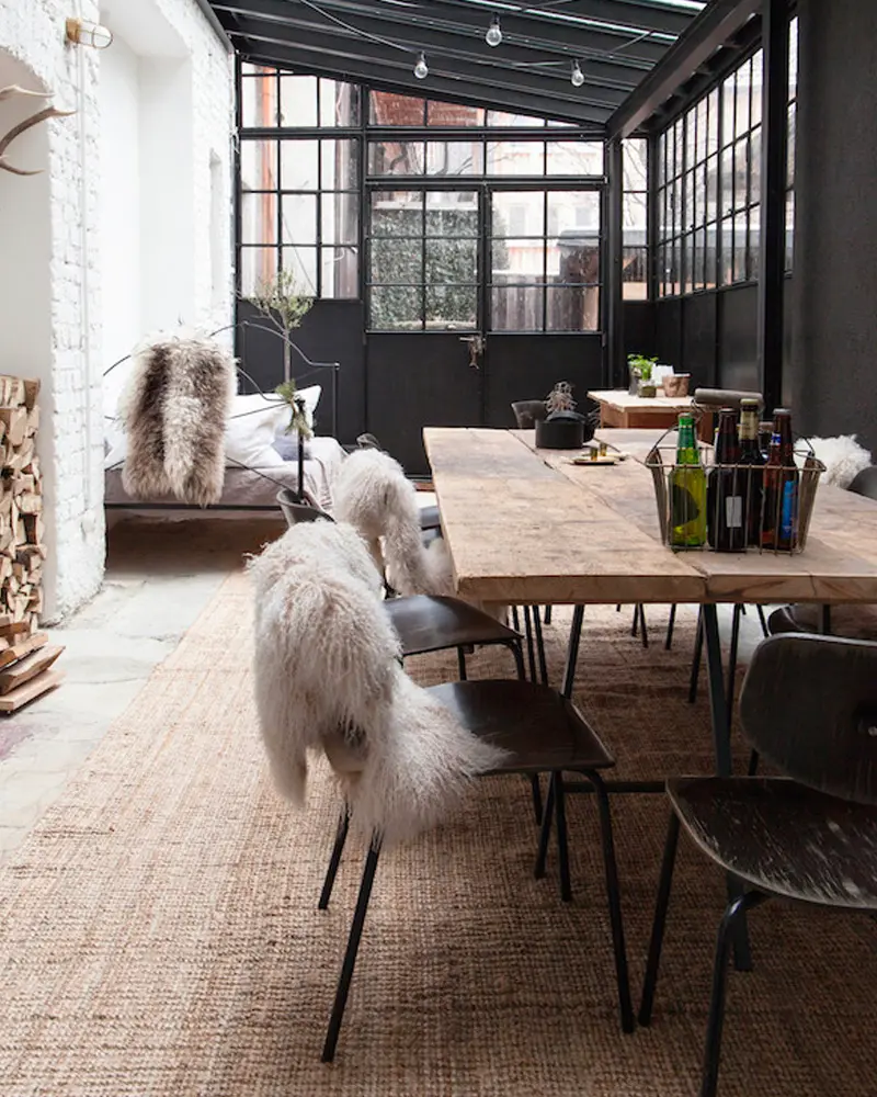 Long wood dining table with sheepskin-covered chairs on a jute rug under a black-framed glass roof