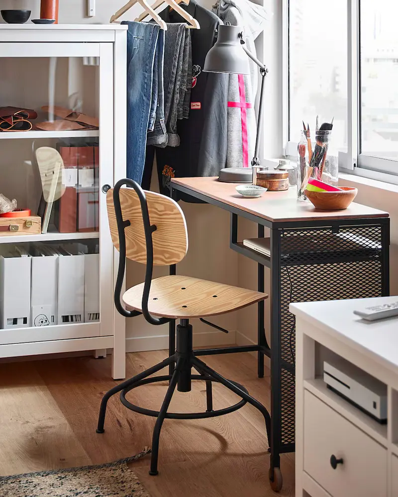 Black metal mesh desk with wood swivel chair, metal task lamp and clothes rack beside a white cabinet