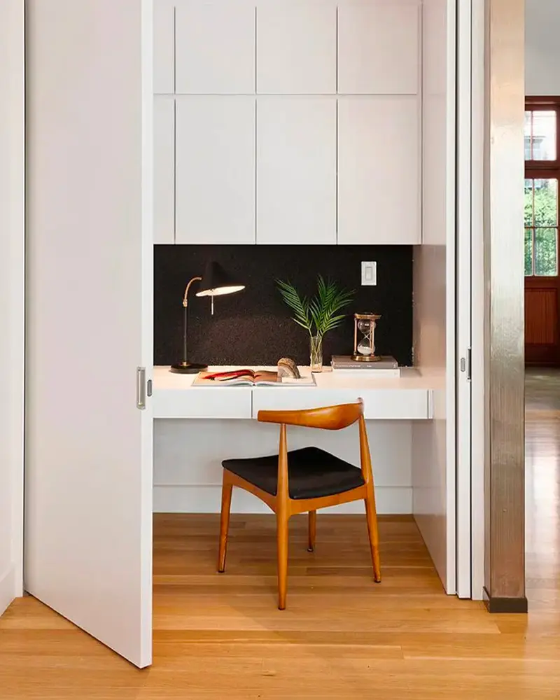 Closet desk with black backsplash, wood chair and small lamp behind open white doors