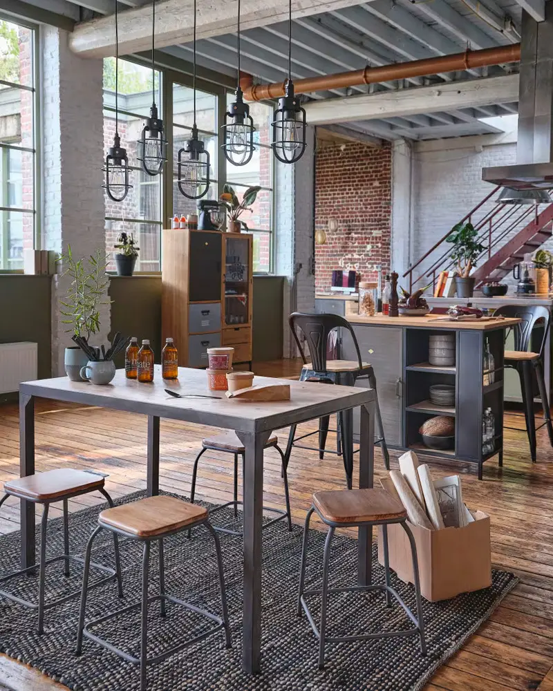 Metal-frame dining table with wood stools and a row of cage pendants in a brick loft