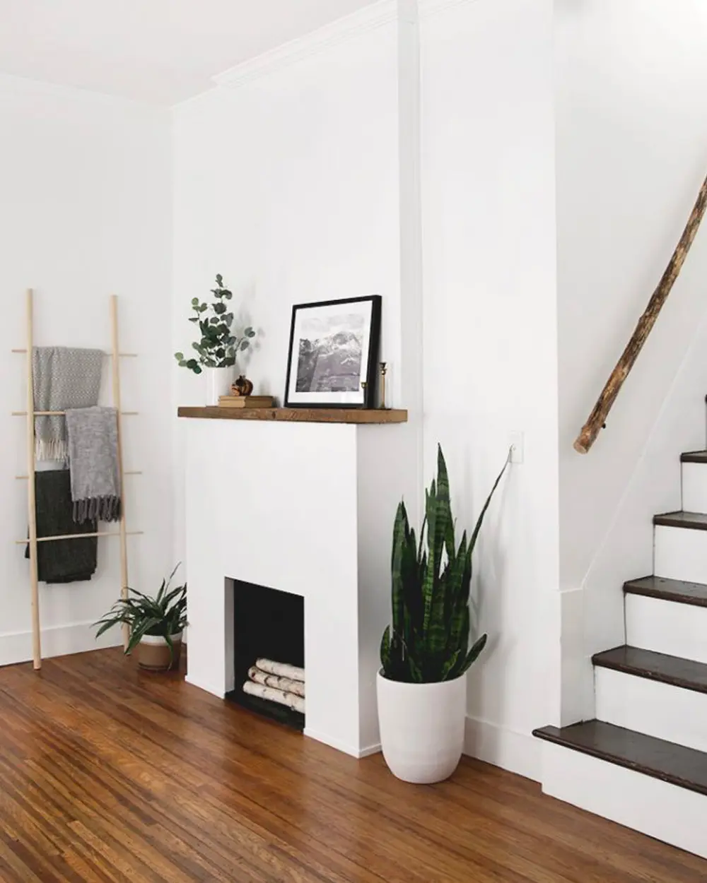 White minimalist faux fireplace with a wood mantel shelf and logs in the opening, next to a staircase
