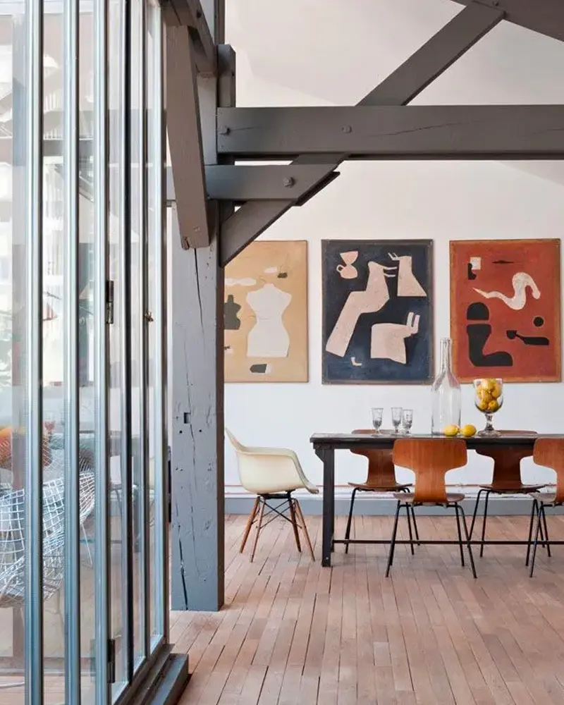 Black dining table with molded plywood chairs, a glass partition, dark beams and abstract prints