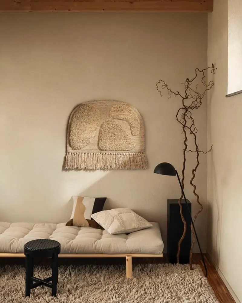 Beige daybed under a fringed woven wall hanging, black side table, branches and shaggy rug