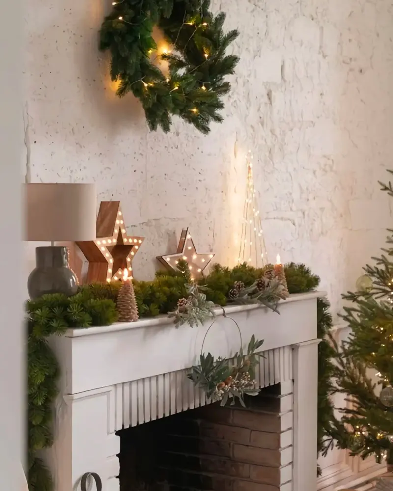 White mantel with a greenery runner, light-up wooden stars, a lit wreath and a white lamp