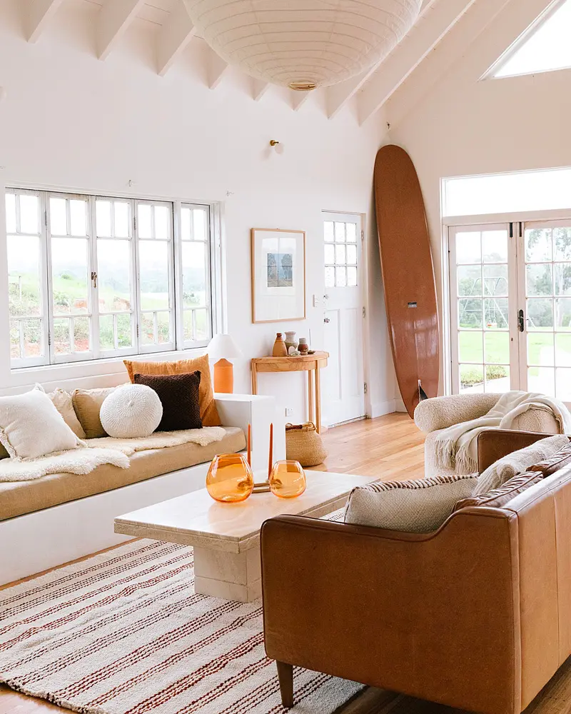 Bright coastal living room with a cognac leather sofa, a white built-in banquette, a wooden surfboard against the wall, a red striped rug and many windows