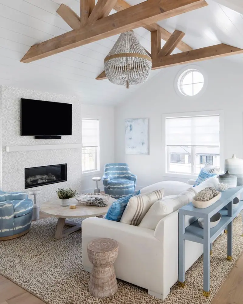 Living room with a vaulted white ceiling and wood beams, a beaded chandelier, blue patterned swivel chairs, a white sofa, a fireplace and a TV