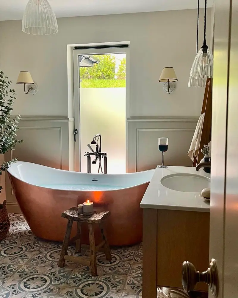 Copper slipper tub on patterned encaustic floor tiles with a small wooden stool, wall sconces and a glass pendant
