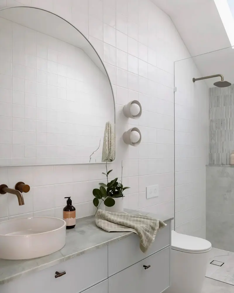 White tiled bathroom with arched mirror, round basin on marble counter and bronze faucet