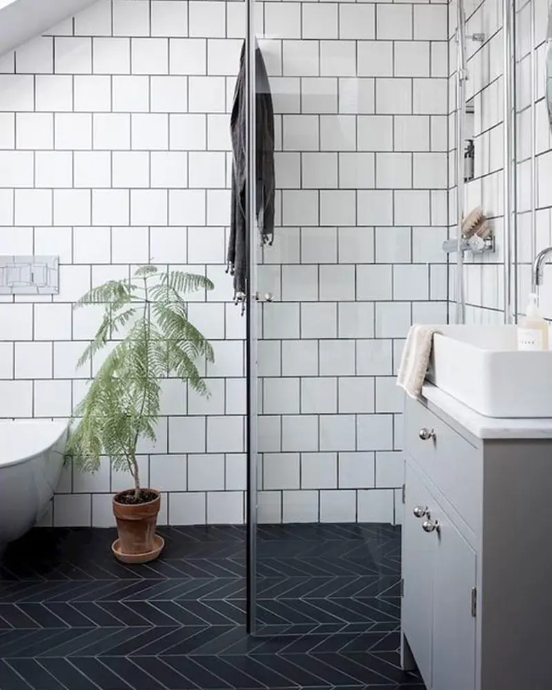 White tiled walk-in shower with black herringbone floor, potted plant and gray vanity