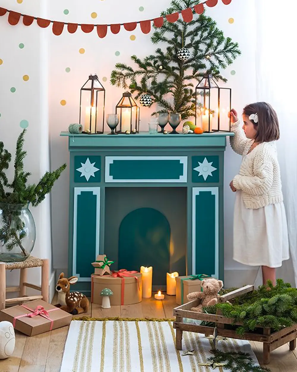 Teal painted cardboard fireplace with white stars, lanterns and a small tree on top, and a child lighting it