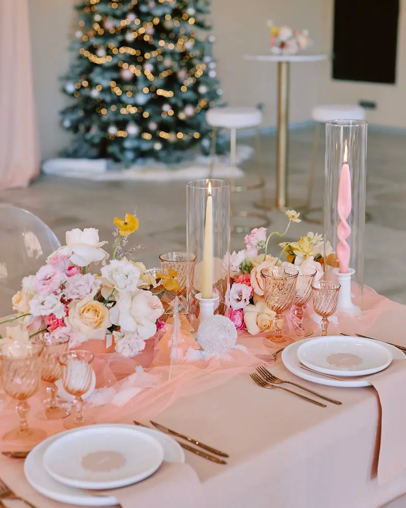 Christmas table with a pale pink runner, white and peach flowers, a pink taper candle in a glass hurricane and white plates