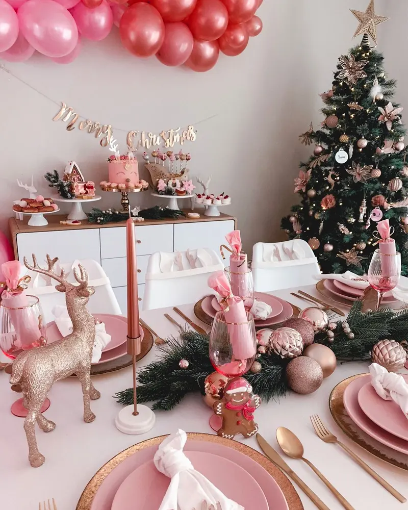 Pink Christmas table with gold reindeer figurine, pink plates, gold flatware and a decorated tree behind